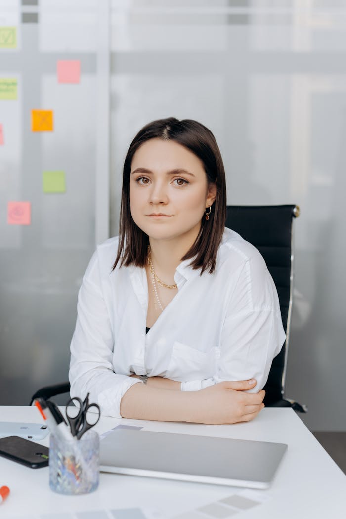 Confident woman in white shirt sitting at desk in a modern office with colorful sticky notes and tech devices.