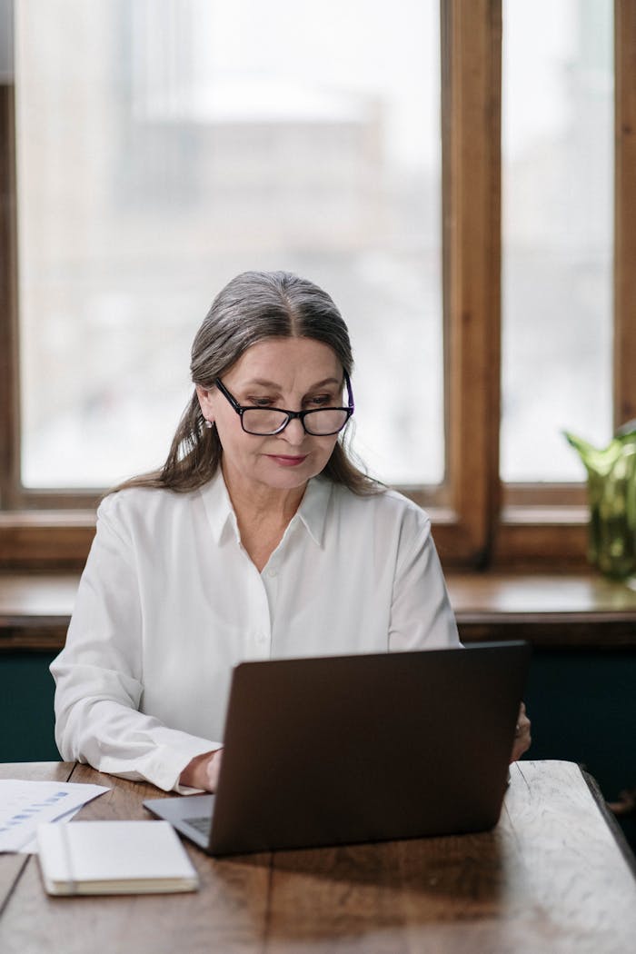 Elderly woman with eyeglasses working on a laptop at home office.